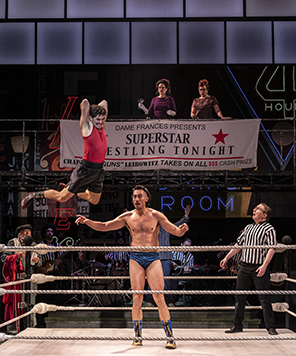 As You Like It, Shakespeare Theatre Company, Wrestling Orlando in red tank t-shirt and black gym shorts flies off the wrestling ring ropes toward Charles in the center of the ring wearing satiny, tight blue shorts. Hanging on the gangway where Dame Frances watches is a bannter, "Superstar Wrestlnig Tonight"